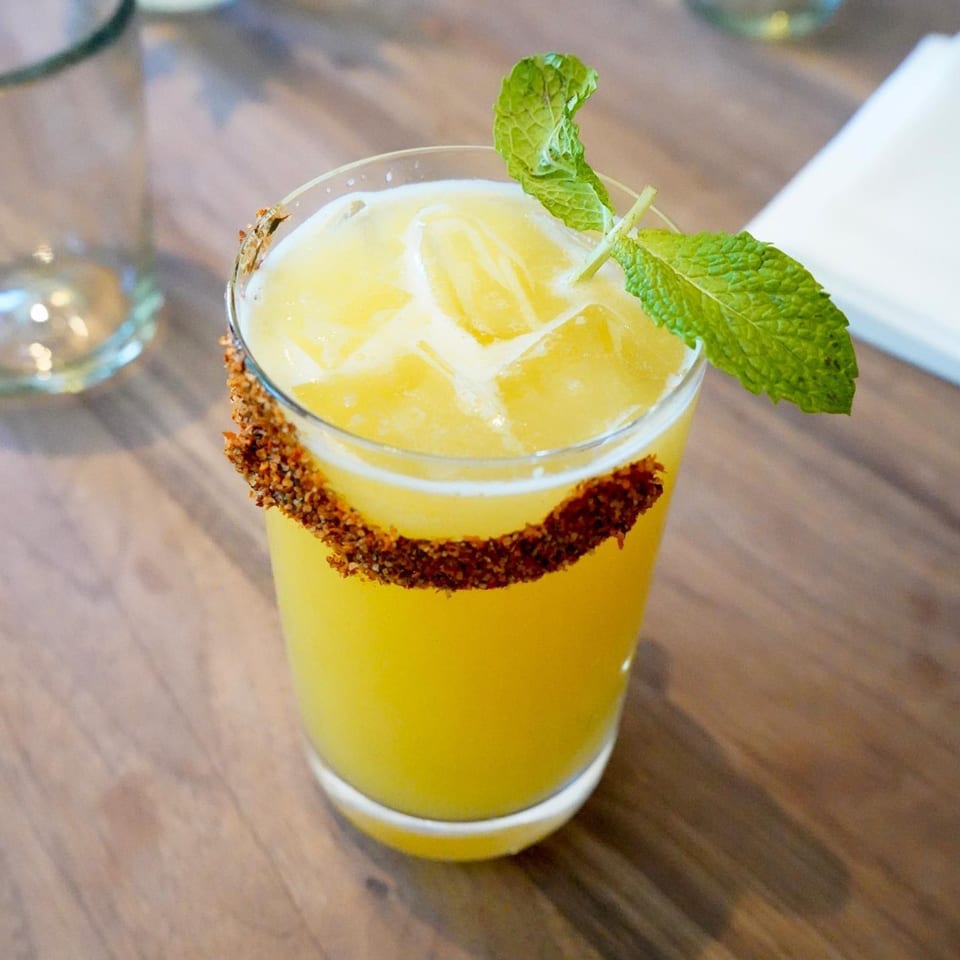 A vibrant yellow cocktail with a chili-lime rim and fresh mint garnish on a wooden table.