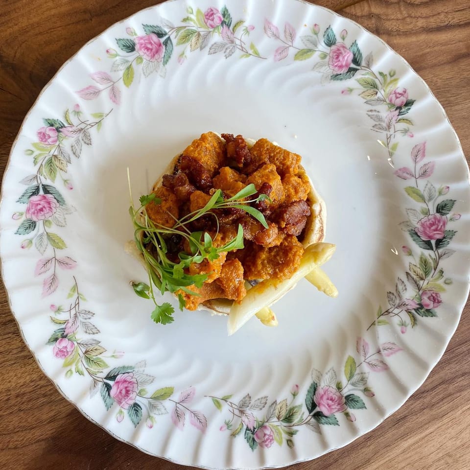 LOLA's Mexican dish with orange-colored meat, microgreens, and white asparagus on a floral plate.