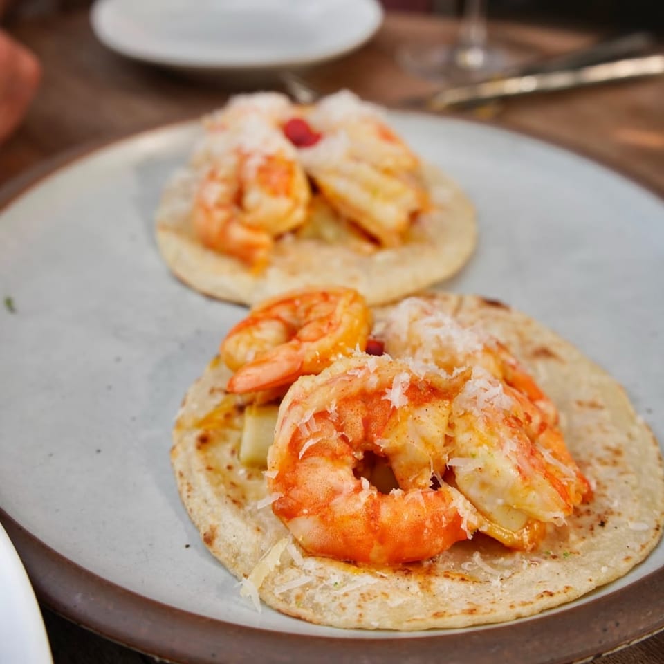 Two LOLA shrimp tacos with grated cheese on corn tortillas, served on a white plate.