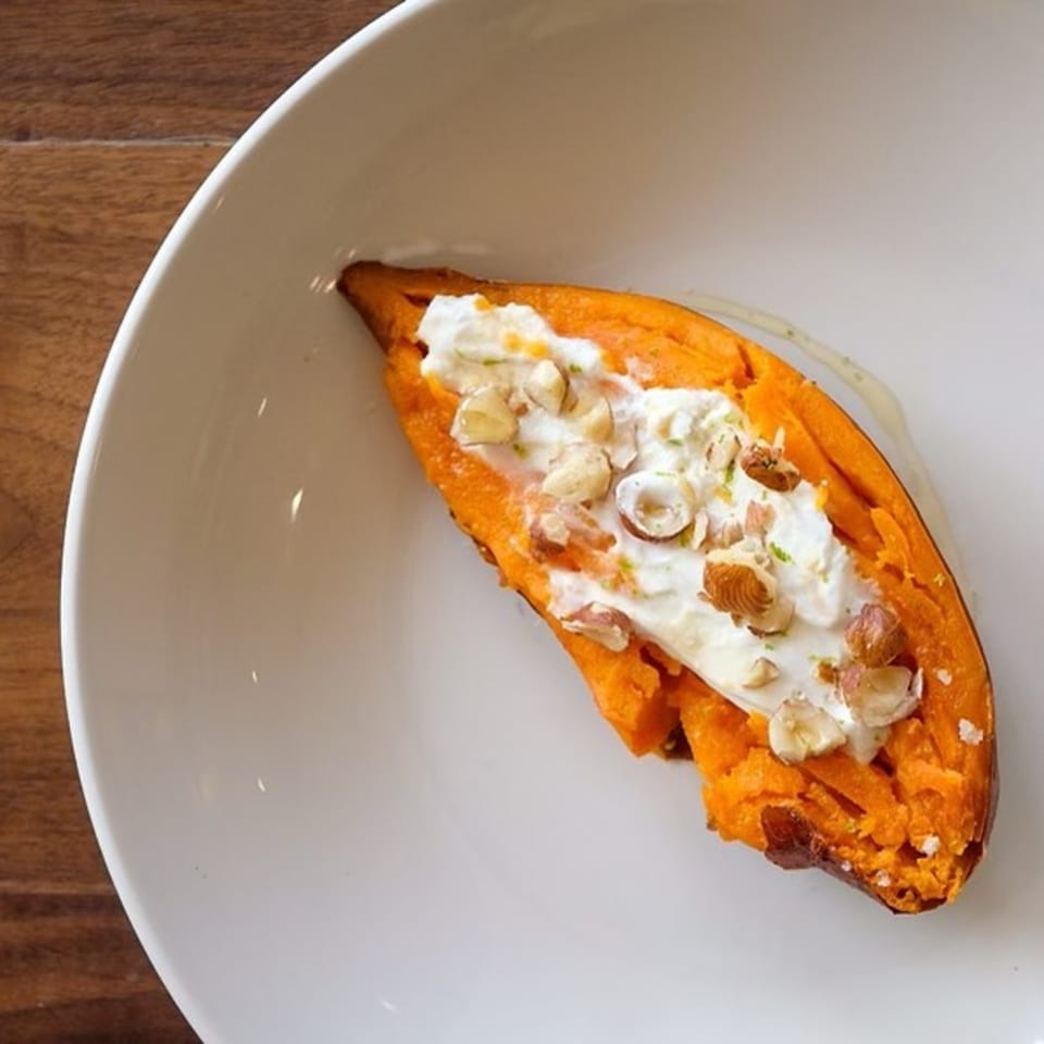 A baked sweet potato topped with white cream and chopped nuts on a white plate.